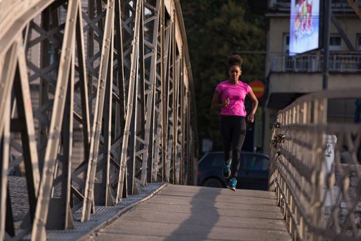 Young sporty african american woman running on sidewalk across the bridge at early morning jogging with city sunrise scene in background