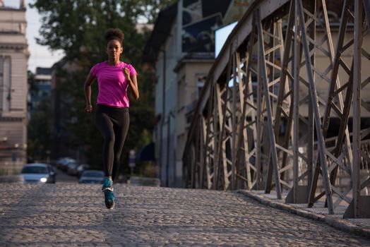 Young sporty african american woman running on sidewalk across the bridge at early morning jogging with city sunrise scene in background