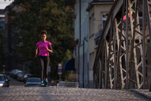 Young sporty african american woman running on sidewalk across the bridge at early morning jogging with city sunrise scene in background