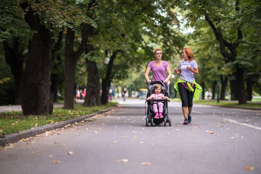 two young healthy women jogging together while pushing a baby stroller at city park