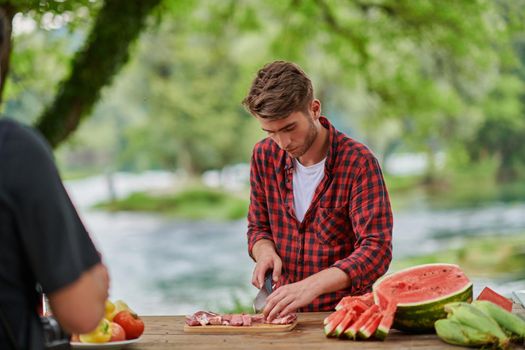 Man cooking tasty food on barbecue grill for outdoor french dinner party near the river on beautiful summer evening in nature