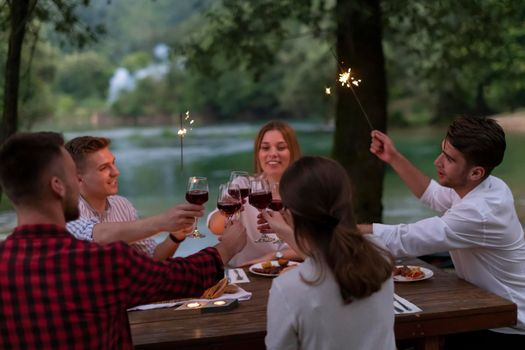 group of happy friends toasting red wine glass while having picnic french dinner party outdoor during summer holiday vacation near the river at beautiful nature