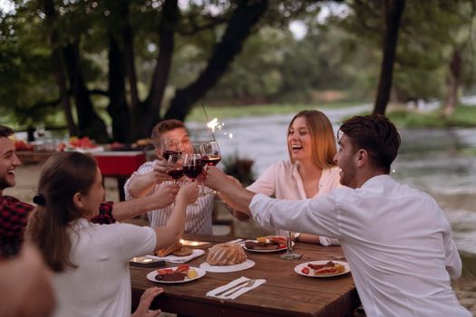 group of happy friends toasting red wine glass while having picnic french dinner party outdoor during summer holiday vacation near the river at beautiful nature