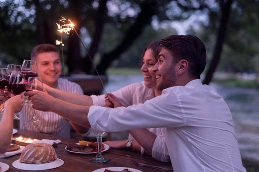 group of happy friends toasting red wine glass while having picnic french dinner party outdoor during summer holiday vacation near the river at beautiful nature