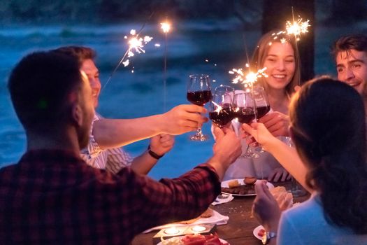 group of happy friends toasting red wine glass while having picnic french dinner party outdoor during summer holiday vacation near the river at beautiful nature