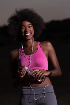 portrait of a young beautiful african american woman with headphones and a bottle of water after jogging in nature beautiful summer night