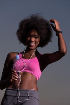 portrait of a young beautiful african american woman with headphones and a bottle of water after jogging in nature beautiful summer night