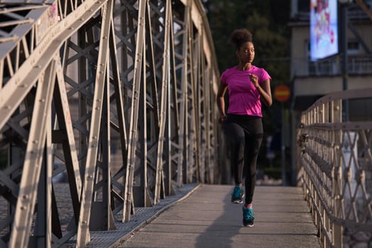 Young sporty african american woman running on sidewalk across the bridge at early morning jogging with city sunrise scene in background