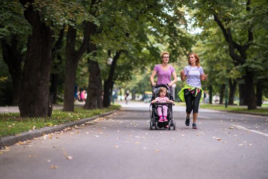 two young healthy women jogging together while pushing a baby stroller at city park