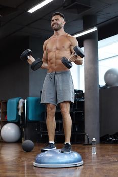 Fit and muscular man working out with bumbbells on gymnastic hemisphere bosu ball in gym