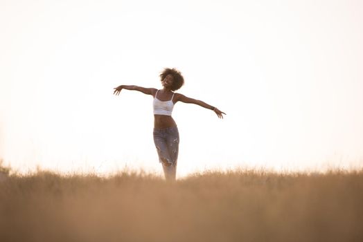 Young beautiful black girl laughs and dances outdoors in a meadow during sunset