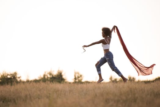 Young beautiful black girl laughs and dances outdoors with a scarf in her hands in a meadow during sunset