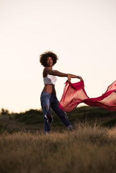 Young beautiful black girl laughs and dances outdoors with a scarf in her hands in a meadow during sunset