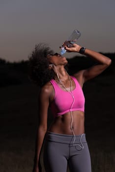 Young beautiful african american woman with headphones drinking water after jogging in nature beautiful summer night