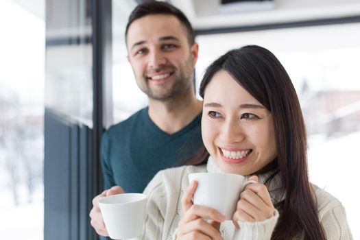 happy young multiethnic couple enjoying morning coffee by the window on cold winter day at home