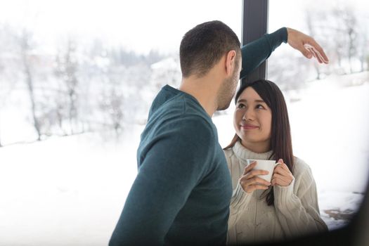 happy young multiethnic couple enjoying morning coffee by the window on cold winter day at home