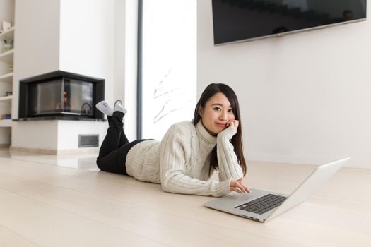 young Asian woman using laptop in front of fireplace on cold winter day at home