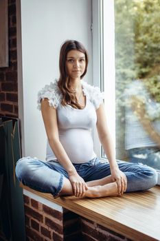 Beautiful pregnant brunette girl sitting on the windowsill. Outside is green, the girl is in white T-shirt and ripped jeans. Looking at the camera