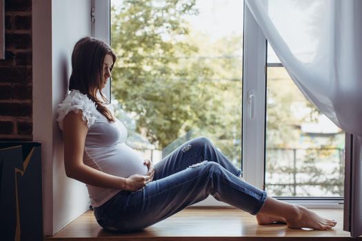 Beautiful pregnant brunette girl sitting on the windowsill. Outside greens, the girl is in white T-shirt and ripped jeans. Looking at the camera