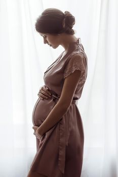 Silhouette of pregnant woman in a pink negligee standing against the window. Side view of happy pregnant woman looking at belly