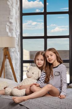 Two little sisters in striped t-shirts are sitting on the sofa in the bright spacious room. They are looking at camera, holding white plush bear