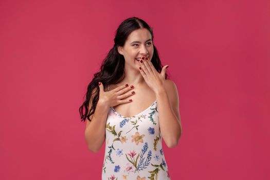 Portrait of a female in a white dress with floral print standing on a pink wall background in studio. She is covering her mouth with hand and looking very happy. People sincere emotions, lifestyle concept. Mockup copy space.