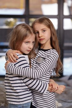 Close up photo of two little sisters in striped t-shirts are sitting on the sofa in the bright spacious room. They hug each other