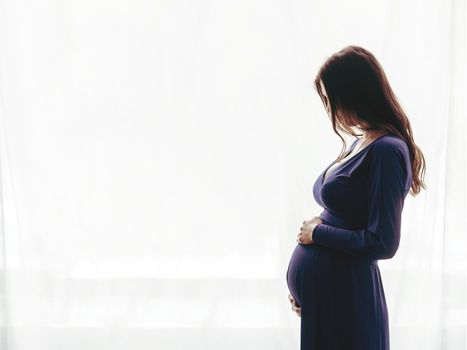 Pregnant woman in a blue dress at home standing against the window. Silhouette of a woman.