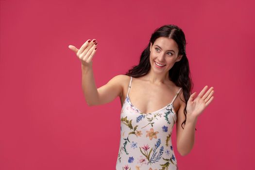 Portrait of a young charming female in a white dress with floral print standing on a pink wall background in studio. She is calling someone and smiling. People sincere emotions, lifestyle concept. Mockup copy space.