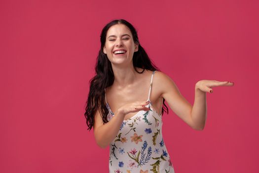 Portrait of a pretty woman in a white dress with floral print standing on a pink wall background in studio. She is laughing and looking happy. People sincere emotions, lifestyle concept. Mockup copy space.