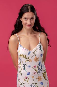 Portrait of a shy pretty woman with a long curly hair, wearing in a white dress with floral print and standing on a pink wall background in studio. People sincere emotions, lifestyle concept. Mockup copy space.