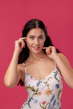 Portrait of a woman with a long curly hair, wearing in a white dress with floral print and standing on a pink wall background in studio. She is touching her face, smiling and looking at the camera. People sincere emotions, lifestyle concept. Mockup copy space.