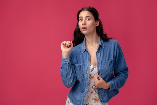 Portrait of a wondered young woman in a white dress with floral print and blue denim shirt standing on a pink wall background in studio. People sincere emotions, lifestyle concept. Mockup copy space.