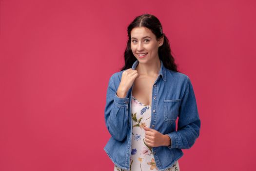Portrait of a happy young woman in a white dress with floral print and blue denim shirt standing on a pink wall background in studio. People sincere emotions, lifestyle concept. Mockup copy space.