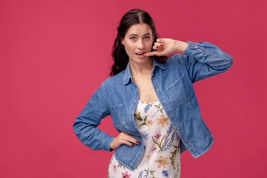 Portrait of an unusual girl in a white dress with floral print and blue denim shirt standing on a pink wall background in studio. She bites her finger and looking at the camera. People sincere emotions, lifestyle concept. Mockup copy space.