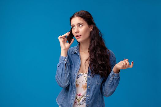 Young brunette woman in a white dress with floral print and blue denim shirt is posing with a smartphone on a blue background. She is calling someone. People sincere emotions, lifestyle concept. Mockup copy space.