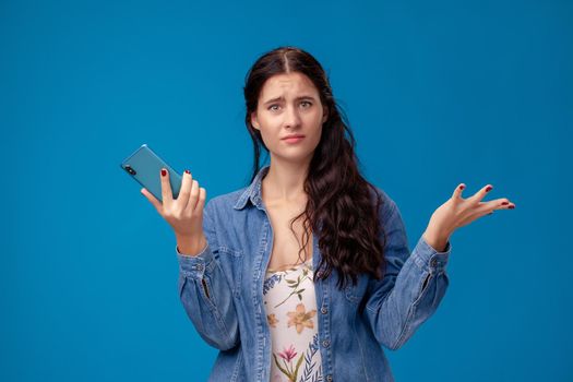 Young beautiful female in a white dress with floral print and blue denim shirt is posing with a smartphone on a blue background. She is looking disappointed. People sincere emotions, lifestyle concept. Mockup copy space.