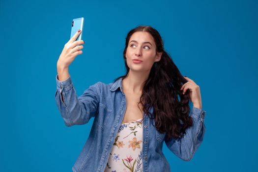 Young pretty female in a white dress with floral print and blue denim shirt is posing with a smartphone on a blue background. She is making a selfie. People sincere emotions, lifestyle concept. Mockup copy space.