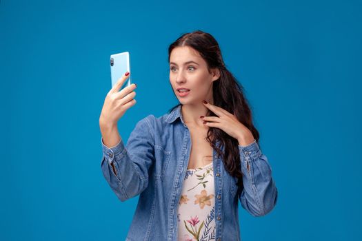 Young charming female in a white dress with floral print and blue denim shirt is posing with a smartphone on a blue background. She is making a selfie. People sincere emotions, lifestyle concept. Mockup copy space.