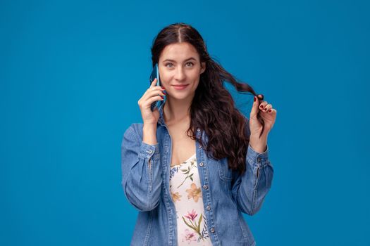 Young attractive woman in a white dress with floral print and blue denim shirt is posing with a smartphone on a blue background. She is talking to someone. People sincere emotions, lifestyle concept. Mockup copy space.
