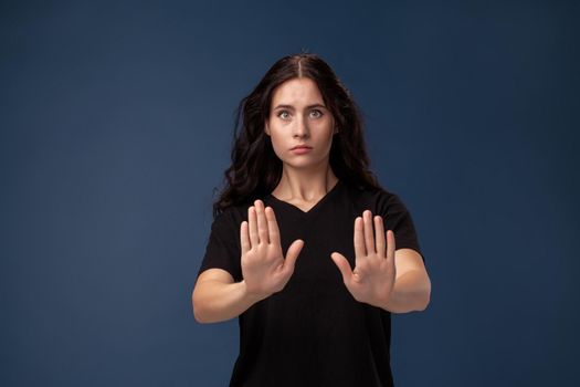 Portrait of disappointed female with curly hair dressed in a black t-shirt. She act like she is asking someone to stop, standing on a gray background.