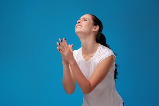 Studio portrait of a young upset woman in white t-shirt against a blue wall background. She act like she is praying. People sincere emotions, lifestyle concept. Mock up copy space.