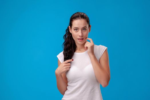 Studio portrait of a brunette woman looking at the camera, dressed in a white t-shirt and standing against a blue wall background. People sincere emotions, lifestyle concept. Mock up copy space.