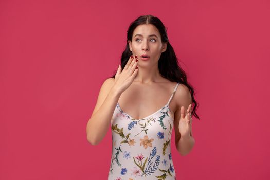 Portrait of a beautiful girl in a white dress with floral print standing on a pink wall background in studio. She is wondered and looking at someone. People sincere emotions, lifestyle concept. Mockup copy space.