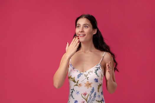 Portrait of a good-looking girl in a white dress with floral print standing on a pink wall background in studio. She is shocked, covering her mouth with her hand and smiling. People sincere emotions, lifestyle concept. Mockup copy space.