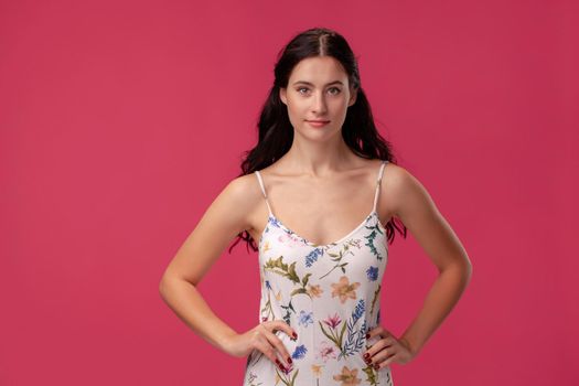 Portrait of a charming brunette woman with a long curly hair, wearing in a white dress with floral print and standing on a pink wall background in studio. She put her hands on her waist and looking at the camera. People sincere emotions, lifestyle concept. Mockup copy space.
