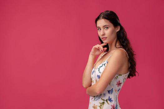 Portrait of a nice brunette woman with a long curly hair, wearing in a white dress with floral print and standing on a pink wall background in studio. She is standing sideways, touches her chin and looking at the camera. People sincere emotions, lifestyle concept. Mockup copy space.