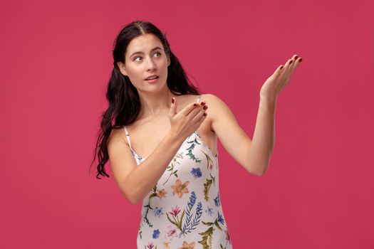 Portrait of a charming lady in a white dress with floral print standing on a pink wall background in studio. She raised her arms up and looking somewhere. People sincere emotions, lifestyle concept. Mockup copy space.