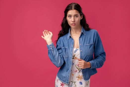 Portrait of a confused young woman in a white dress with floral print and blue denim shirt standing on a pink wall background in studio. People sincere emotions, lifestyle concept. Mockup copy space.