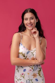 Portrait of a good-looking brunette woman with a long curly hair, wearing in a white dress with floral print and standing on a pink wall background in studio. She touches her chin and smiling at the camera. People sincere emotions, lifestyle concept. Mockup copy space.